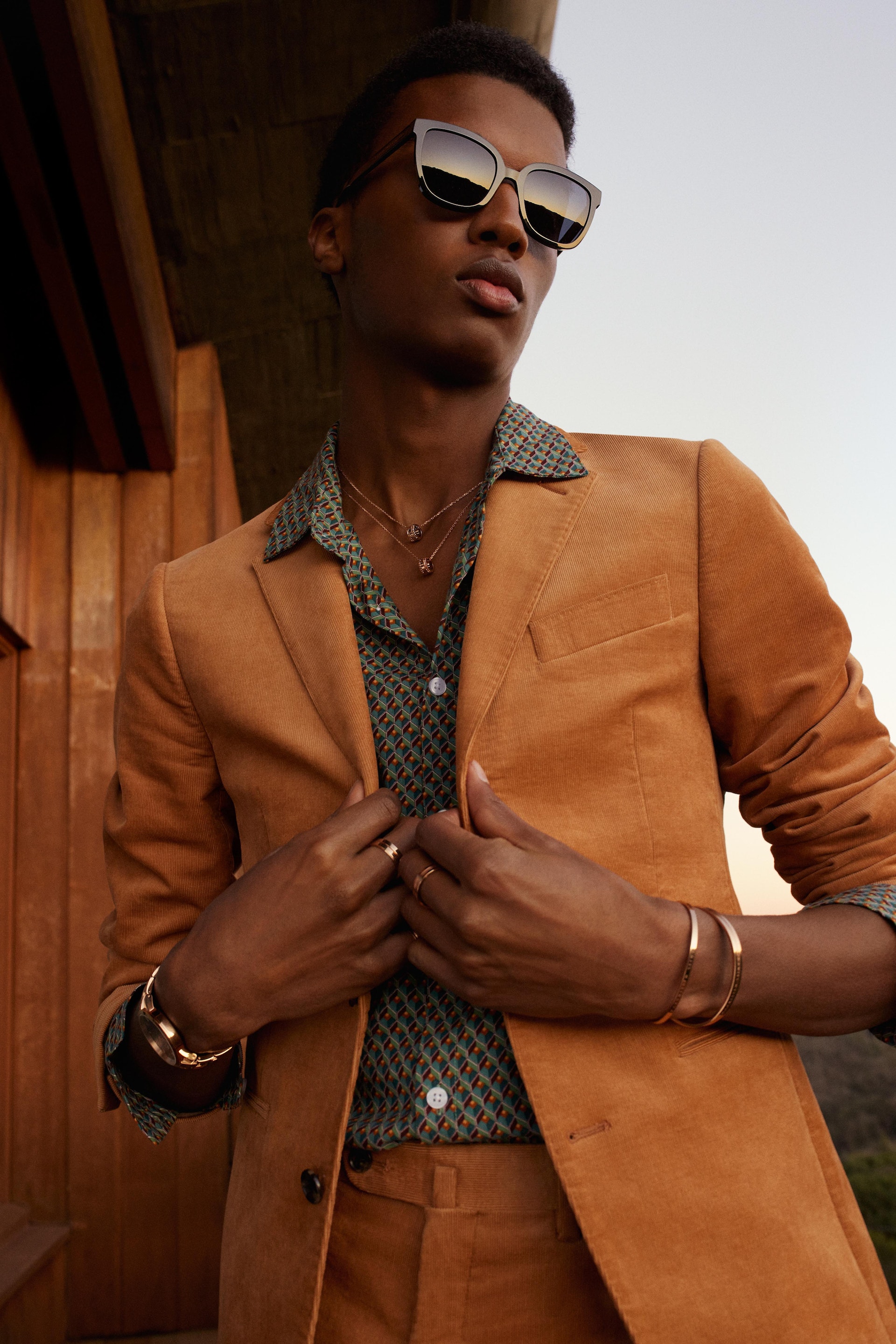 Stylish African-American male model poses outdoors wearing sunglasses, suit, patterned shirt, necklace, bracelets, and rings against a neutral background.