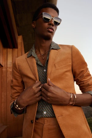 Stylish African-American male model poses outdoors wearing sunglasses, suit, patterned shirt, necklace, bracelets, and rings against a neutral background.