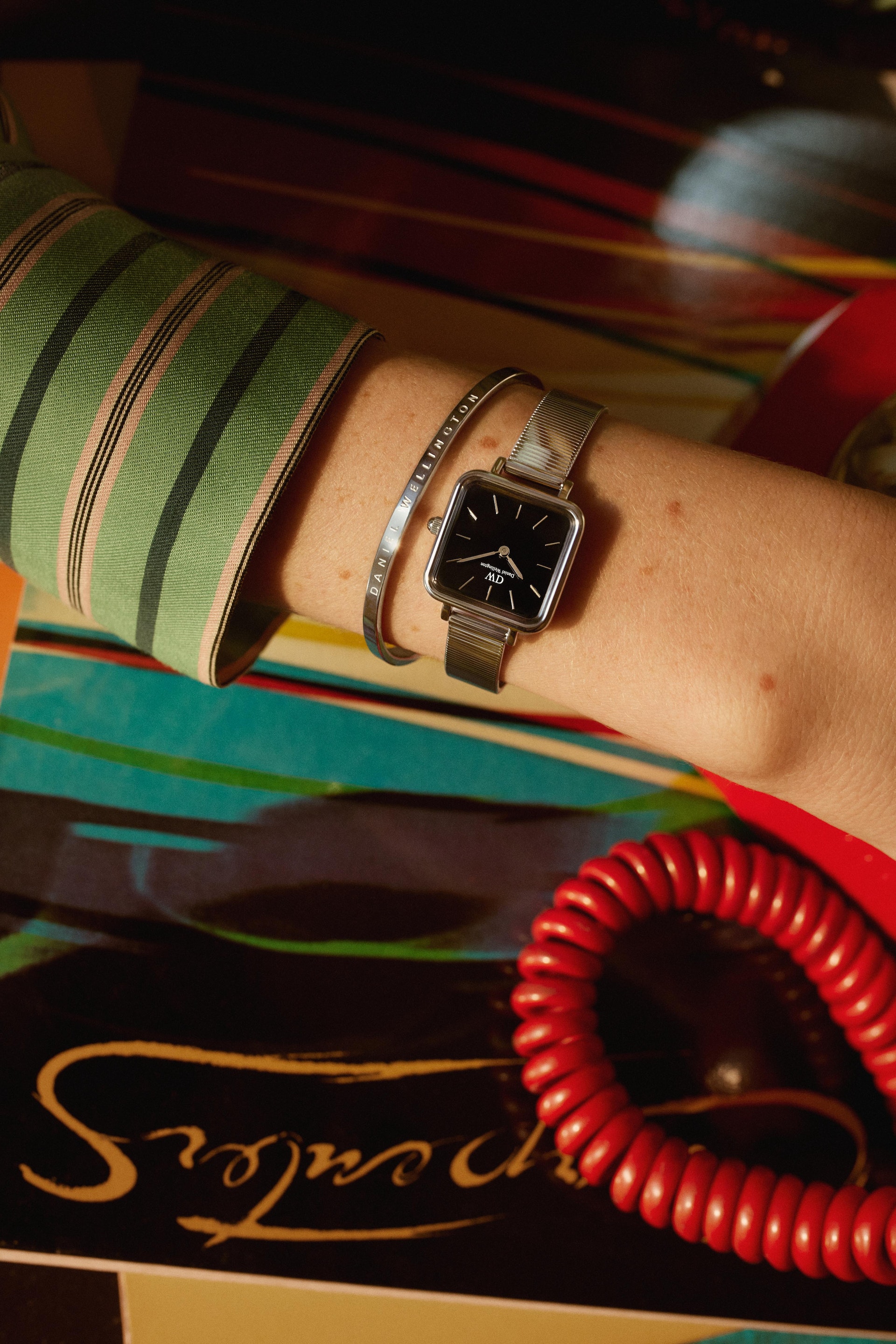 Close-up of a wrist with a silver wristwatch with a black face and a silver bracelet, set against a colorful patterned background.