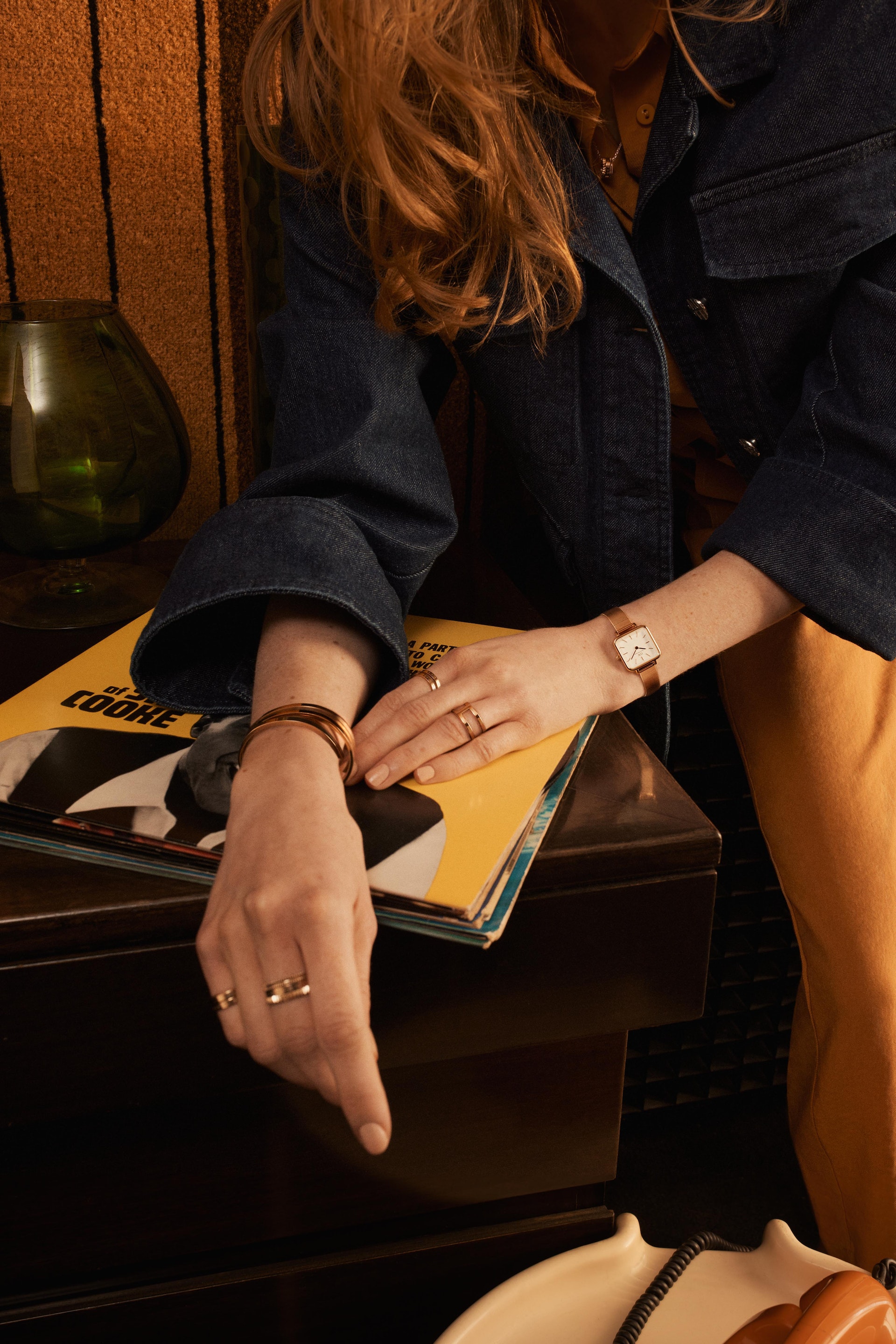 Blonde woman in a denim jacket poses with jewelry, vintage vinyl records, and home decor in warm studio lighting for a fashion editorial.