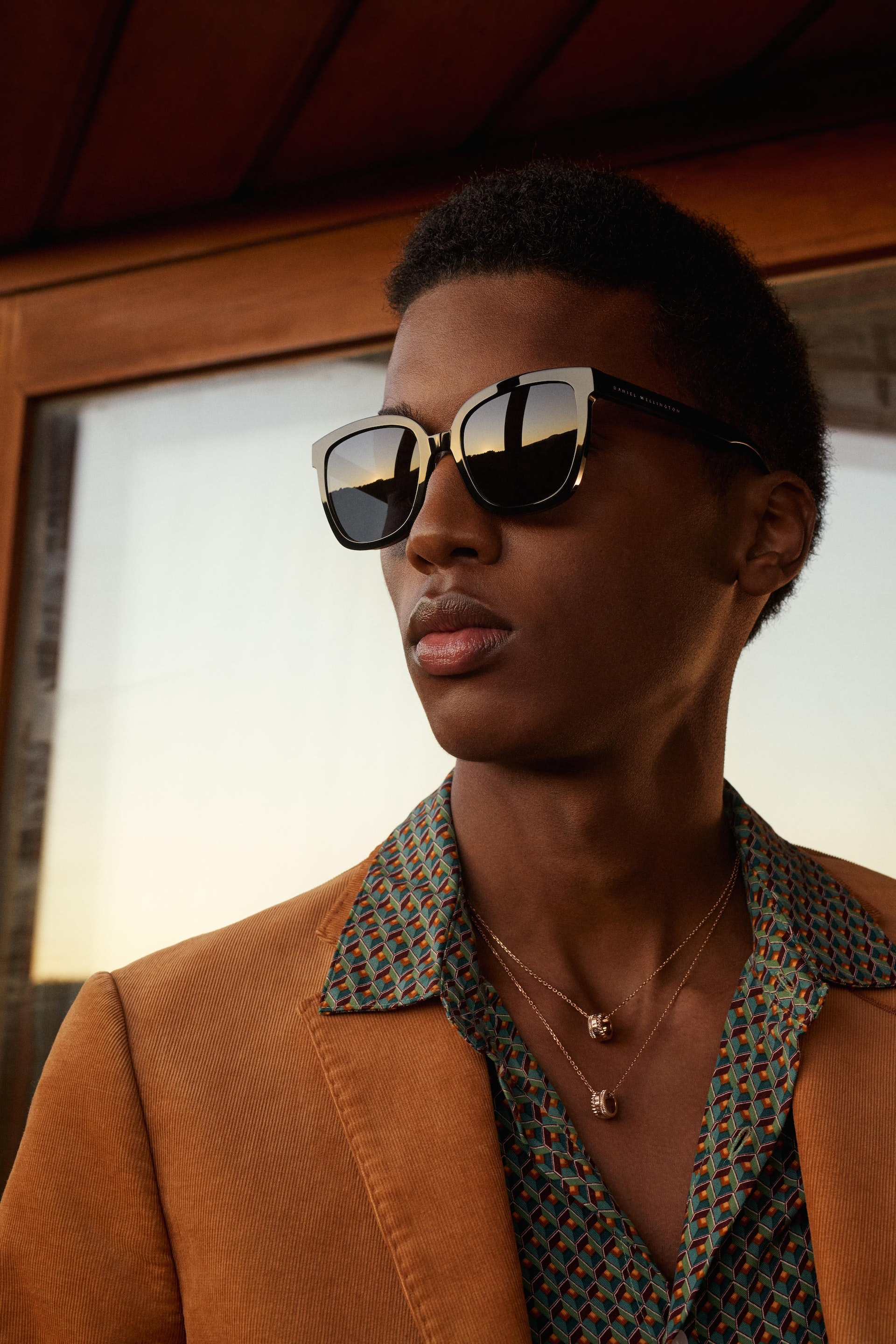Close-up of a Black male model in an orange blazer and patterned shirt, wearing sunglasses and necklaces, against a neutral background.
