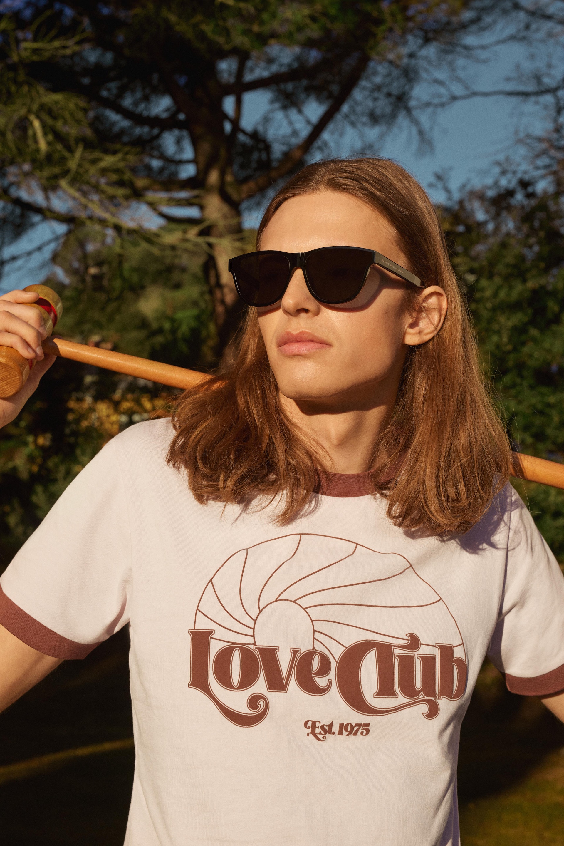 Adult male with long hair and sunglasses stands outdoors holding a sports mallet, wearing a white t-shirt with a graphic print.