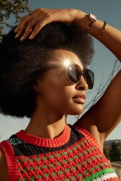 Black woman wearing sunglasses and a patterned knit top with sun glare on the lens outdoors against a clear sky.