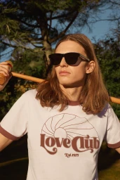 Model wearing dark sunglasses and white ringer t-shirt holding wooden pole outdoors in a park.