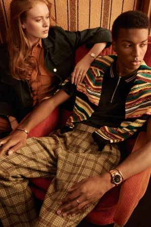 A man and a woman seated together wearing patterned clothing and watches against a textured backdrop