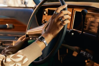 Close-up of a person's hands wearing a silver mesh-strap watch and rings holding a vintage car steering wheel.