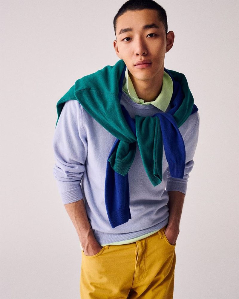 Portrait of an Asian man wearing casual fashion: a light-blue sweater, yellow pants, and a green sweater draped over his shoulders.