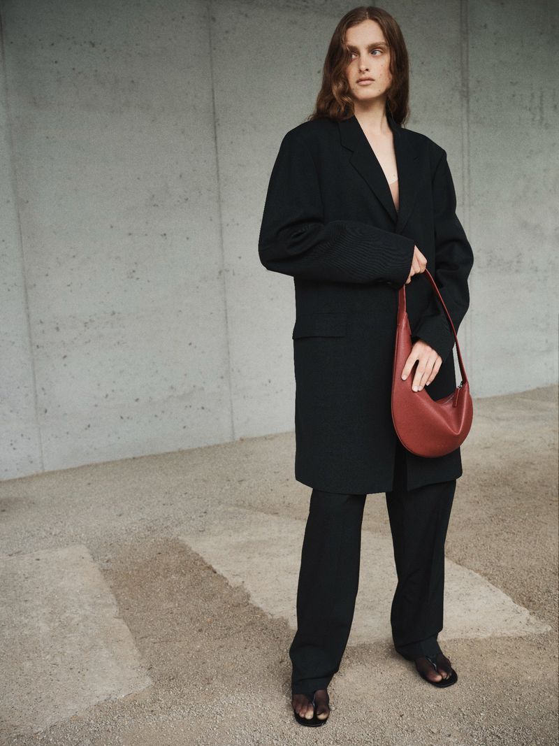 A woman poses in an oversized black blazer and pants, holding a red crescent bag, against a gray concrete wall, in a fashion editorial.