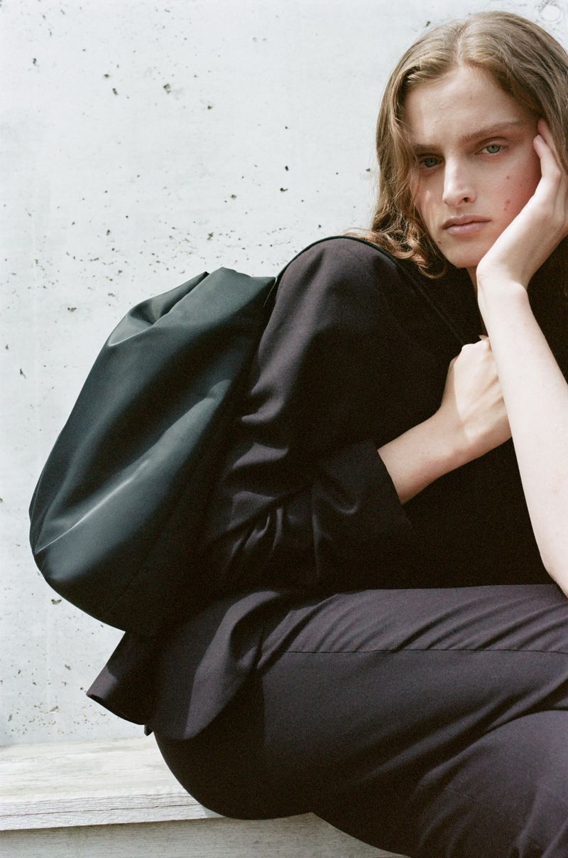 Fashion image of a woman in a black suit and green bag sitting against a concrete wall looking off camera with a thoughtful expression.