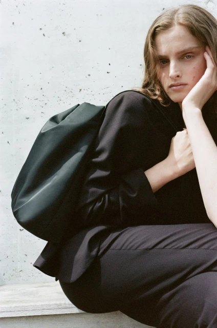 Fashion image of a woman in a black suit and green bag sitting against a concrete wall looking off camera with a thoughtful expression.