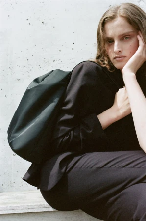 Fashion image of a woman in a black suit and green bag sitting against a concrete wall looking off camera with a thoughtful expression.