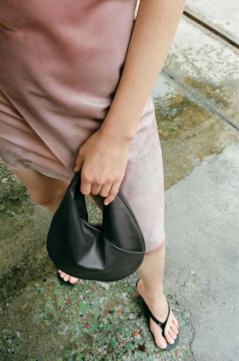 Woman in a pink dress holds a black purse while standing on a concrete surface, creating a fashion and lifestyle composition.