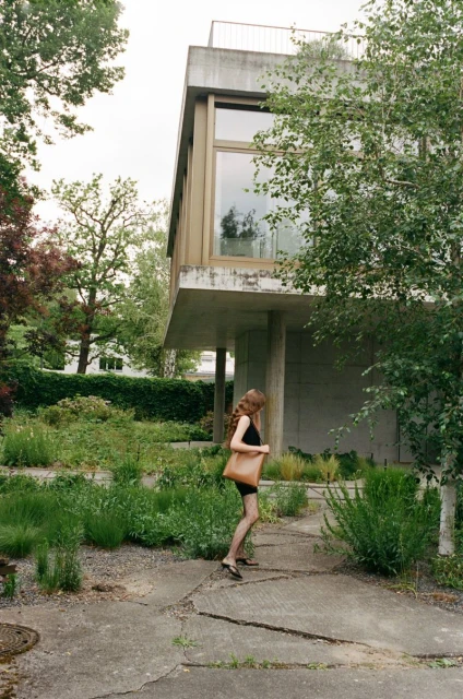 Adult female walking near a modern concrete house surrounded by greenery, carrying a brown leather bag on a sunny day.