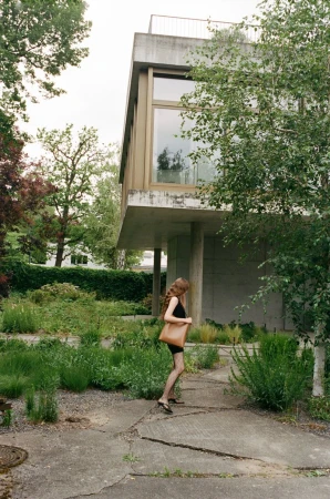 Adult female walking near a modern concrete house surrounded by greenery, carrying a brown leather bag on a sunny day.