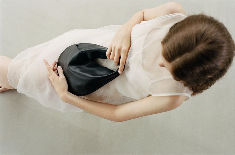 Model in a cream sleeveless dress holding a black leather handbag against a grey seamless background