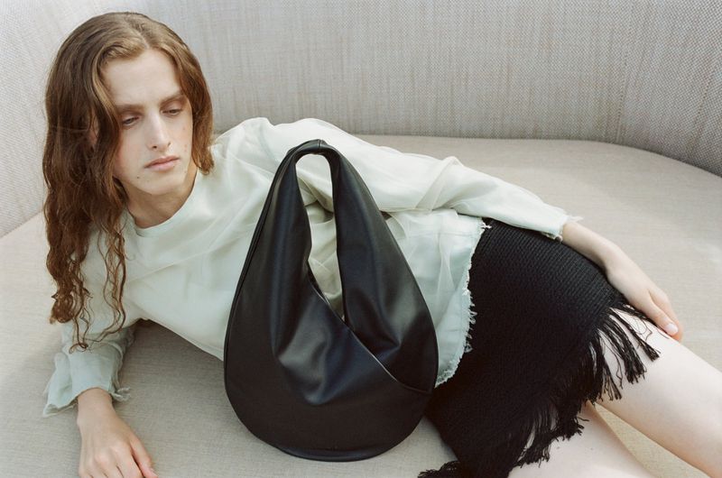 Fashion photo of a reclining adult with a stylish black leather handbag on a light beige couch in a soft indoor light setting.
