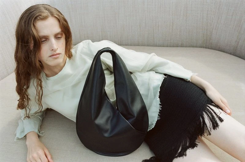 Fashion photo of a reclining adult with a stylish black leather handbag on a light beige couch in a soft indoor light setting.