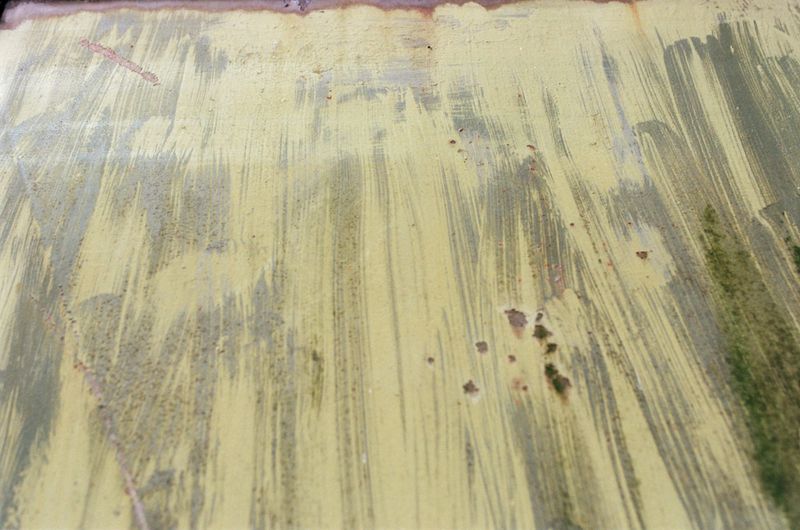 Close-up of an abstract surface, showing a blend of faded yellow and green paint with brushstroke textures and scattered imperfections.