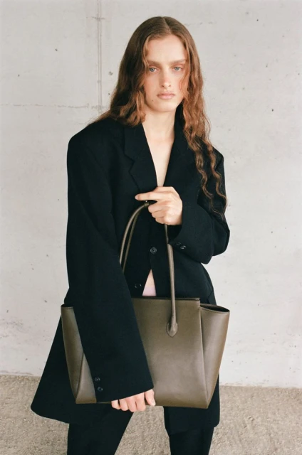 Fashion shot of a young adult woman with long wavy hair wearing a black blazer and holding a taupe leather tote bag indoors.