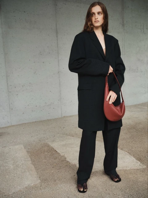 Model in black blazer holding a reddish-brown leather handbag against a concrete wall and stone ground.