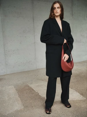 Model in black blazer holding a reddish-brown leather handbag against a concrete wall and stone ground.