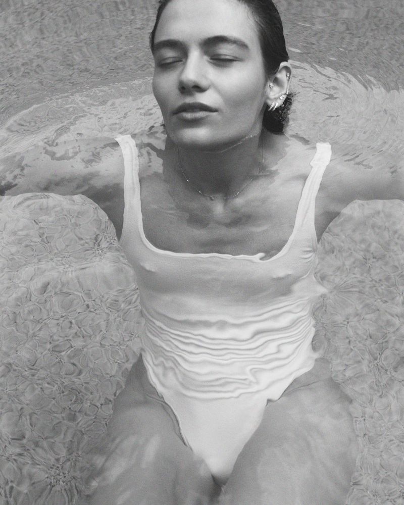 Woman in a white swimsuit floats in water, eyes closed, with light reflecting off the surface in a black and white portrait.