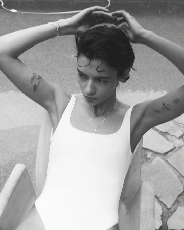 Monochrome portrait of a young adult female with short hair wearing a white swimsuit, sitting outside with her hands behind her head.