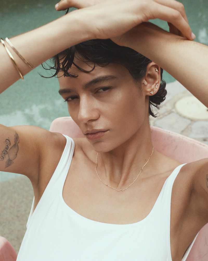 Adult woman in a white tank top poses outdoors with a turquoise pool in the background. She wears minimal jewelry.