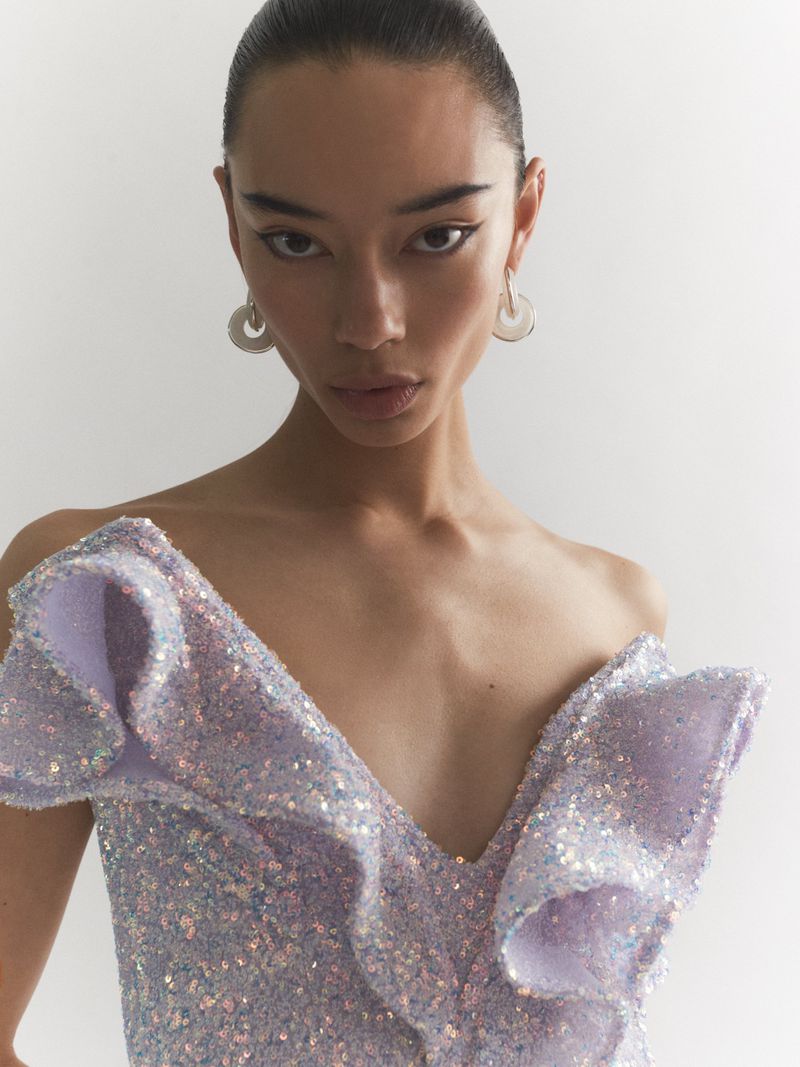 Close-up of an adult female model in a studio setting, wearing a sequined lavender dress with ruffles and hoop earrings, looking at camera.