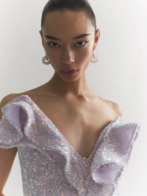 Close-up of an adult female model in a studio setting, wearing a sequined lavender dress with ruffles and hoop earrings, looking at camera.