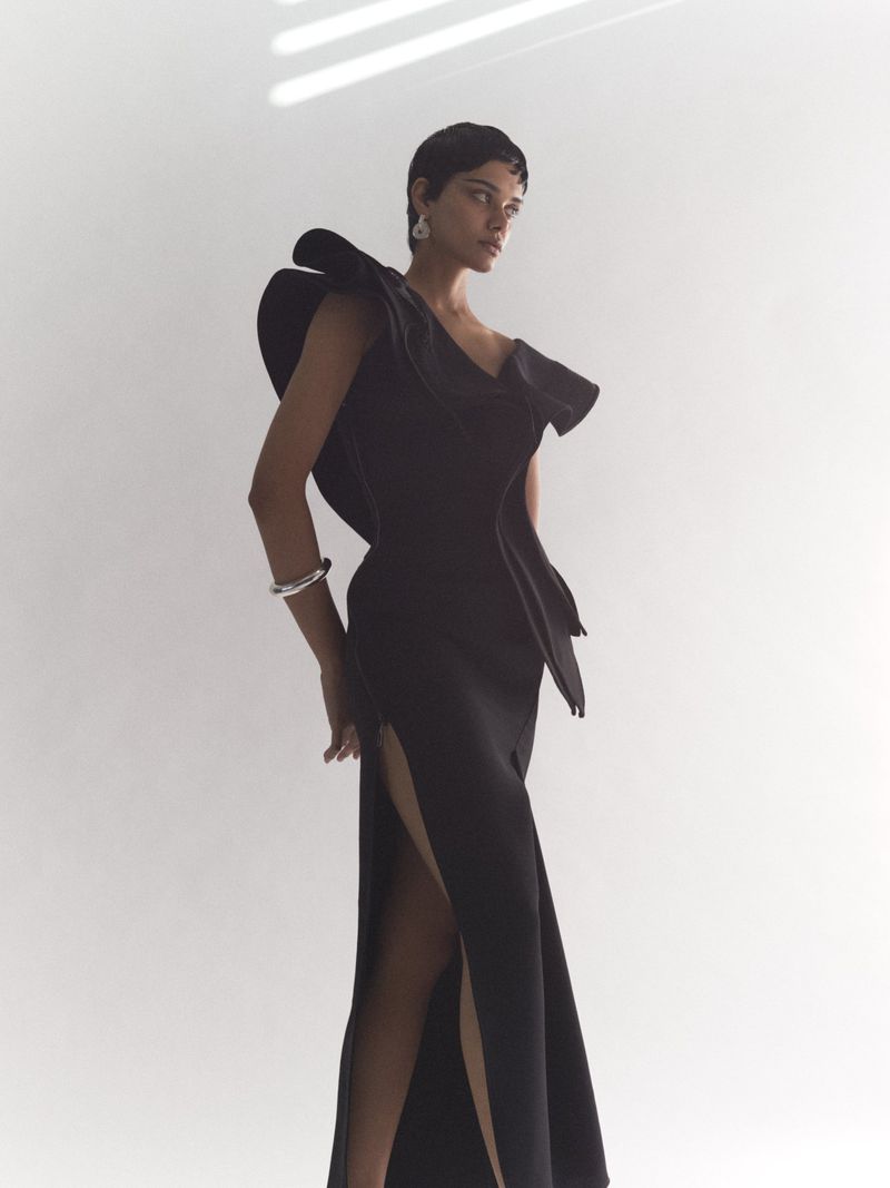 Elegant woman poses in a sophisticated black gown with a dramatic shoulder accent, paired with silver bracelet and earrings, on white backdrop.