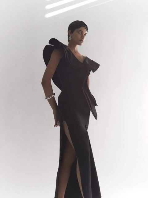 Elegant woman poses in a sophisticated black gown with a dramatic shoulder accent, paired with silver bracelet and earrings, on white backdrop.