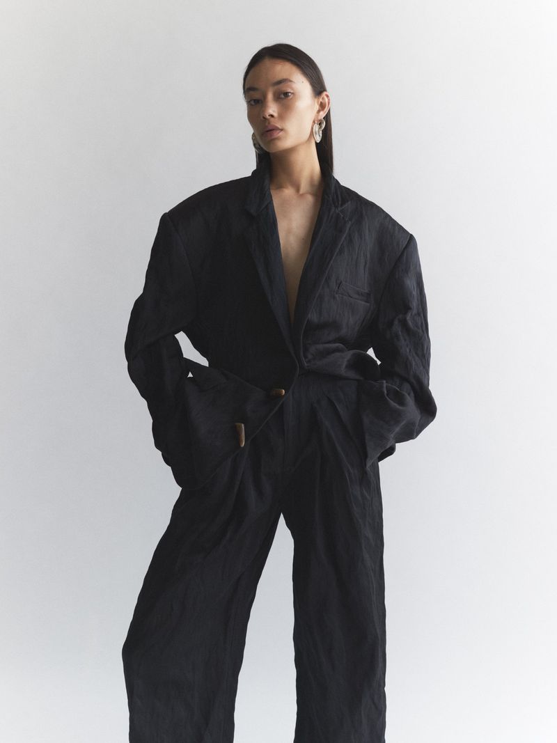 Woman poses in a textured black blazer and pants against a white backdrop. Studio lighting emphasizes the fabric and form.