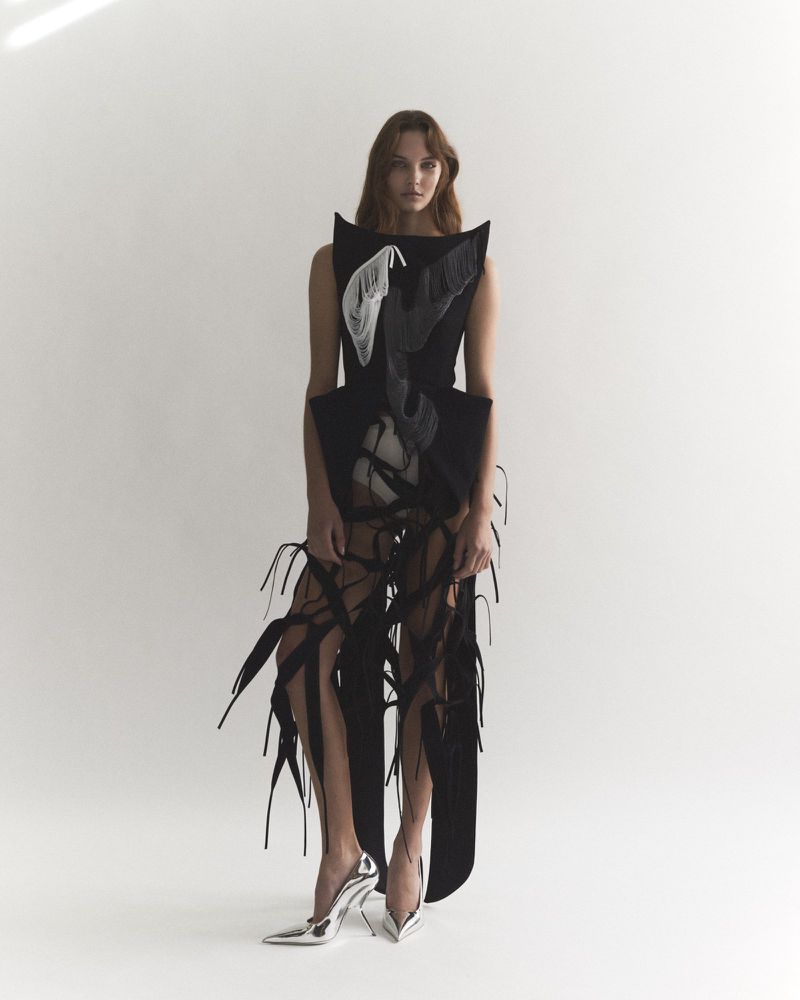 A fair-skinned woman with long brown hair is posing in a sleeveless black avant-garde dress and silver high heels against a white backdrop.