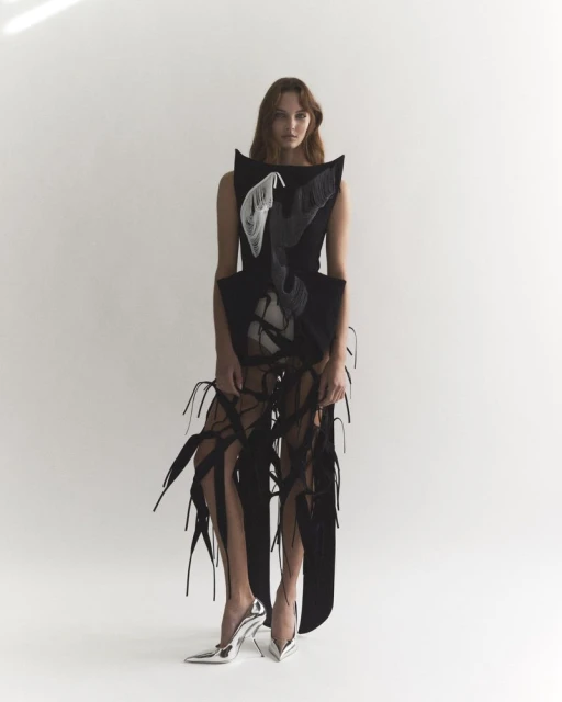 A fair-skinned woman with long brown hair is posing in a sleeveless black avant-garde dress and silver high heels against a white backdrop.