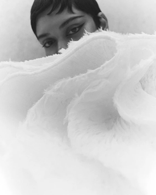 Close-up studio shot of a young adult female model peeking above a ruffled white textile, displaying dark eye makeup against a soft, neutral backdrop.