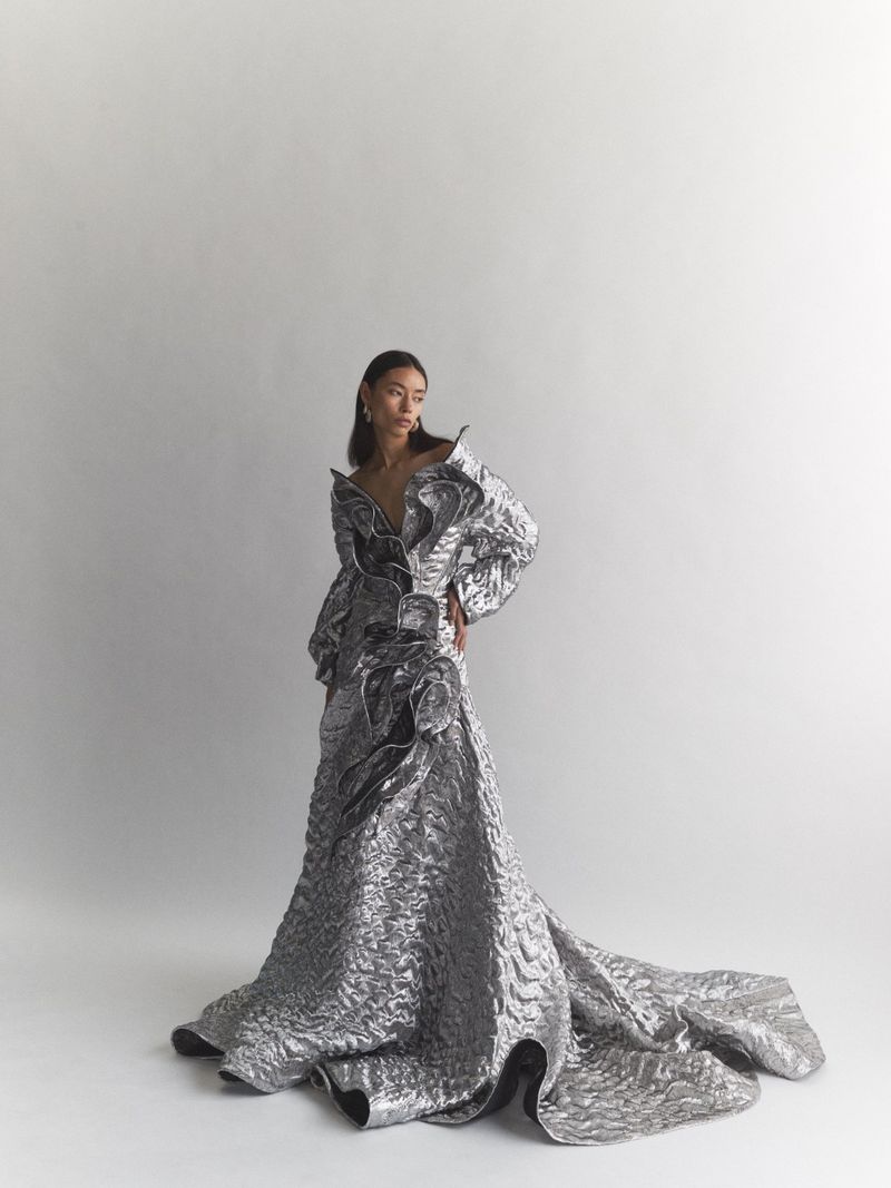 Woman poses in shimmering silver gown with dramatic ruffles and a long train against a bright studio backdrop. Fashion editorial shot.