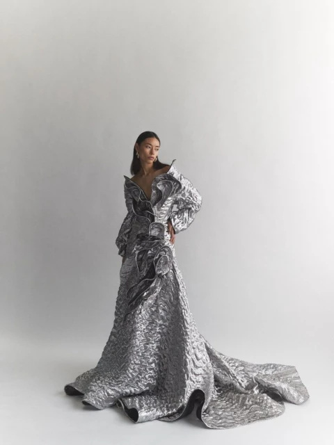 Woman poses in shimmering silver gown with dramatic ruffles and a long train against a bright studio backdrop. Fashion editorial shot.