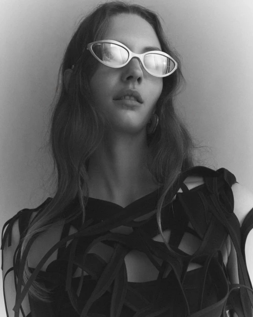 Monochrome portrait of a woman wearing reflective sunglasses and a dark, intricately designed top against a light background.