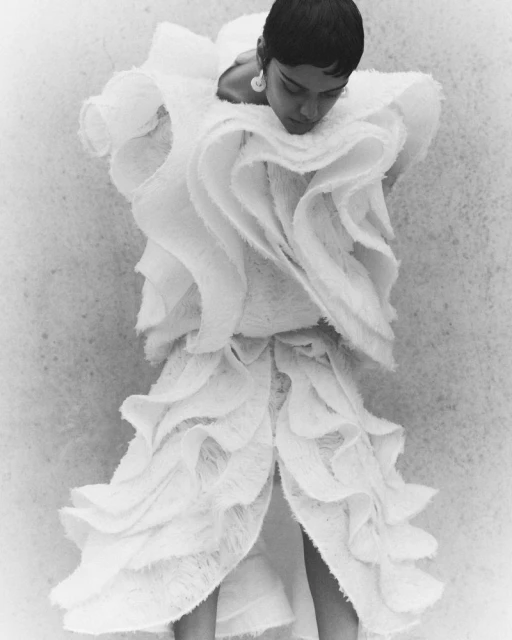 Woman in a white, ruffled, avant-garde dress against a textured grey background. Close-up view of a high fashion editorial.