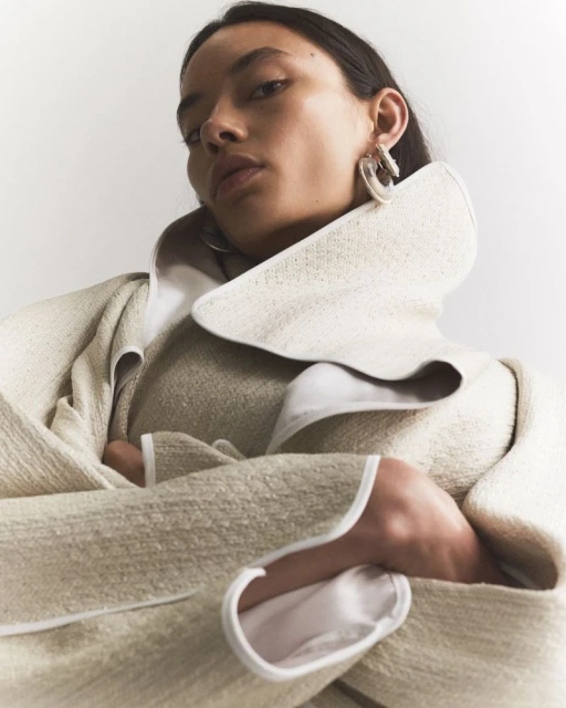 Fashion shot of a woman in a stylish beige wool coat with wide lapels, posing indoors against a bright white backdrop, showcasing contemporary design.