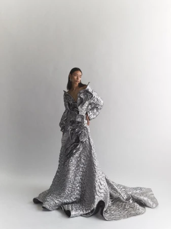 Model wearing a metallic textured long gown with dramatic ruffles standing against a clean white seamless studio background.