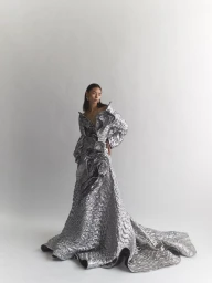 Model wearing a metallic textured long gown with dramatic ruffles standing against a clean white seamless studio background.