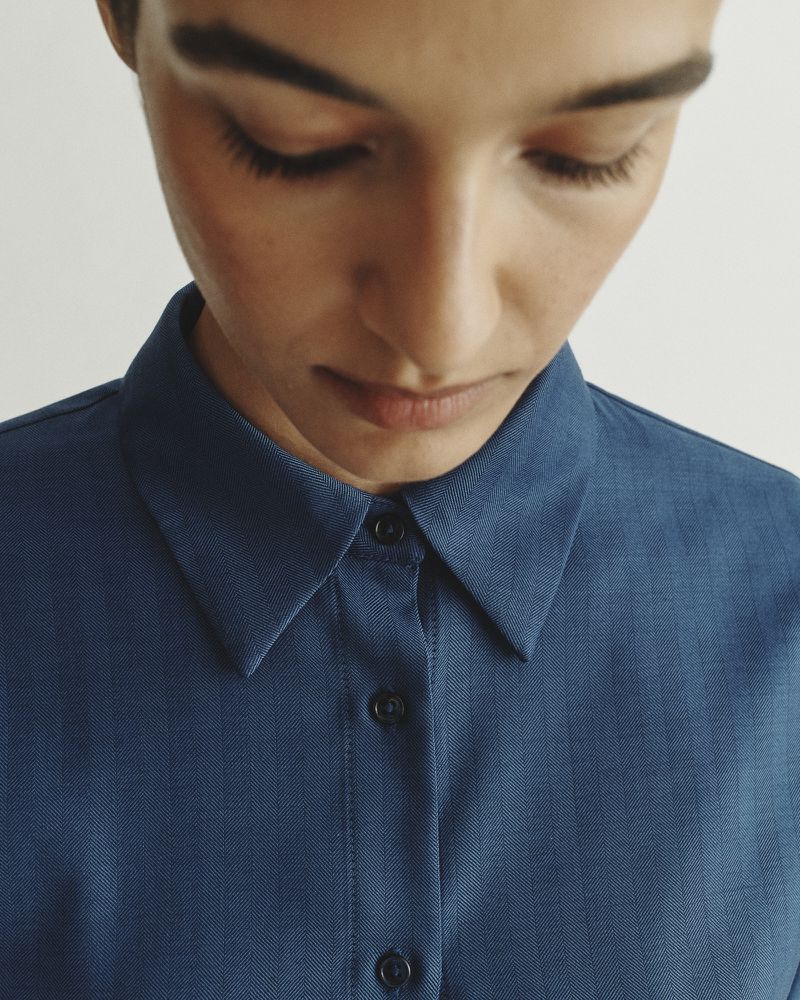Close-up of an adult female in a blue button-down shirt, looking down, emphasizing minimalist fashion and neutral background.