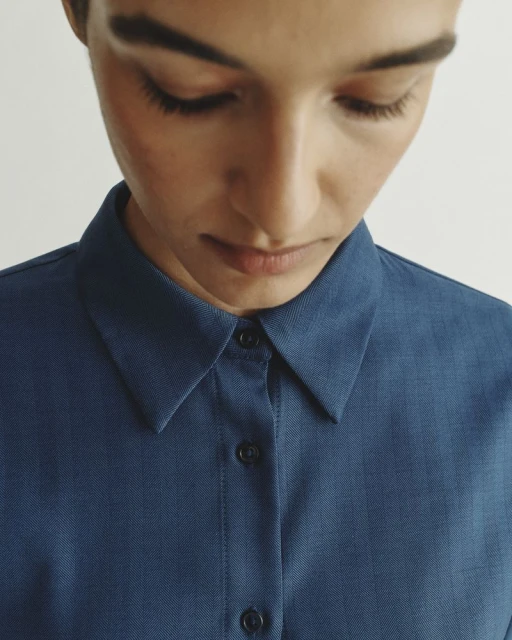 Close-up of an adult female in a blue button-down shirt, looking down, emphasizing minimalist fashion and neutral background.