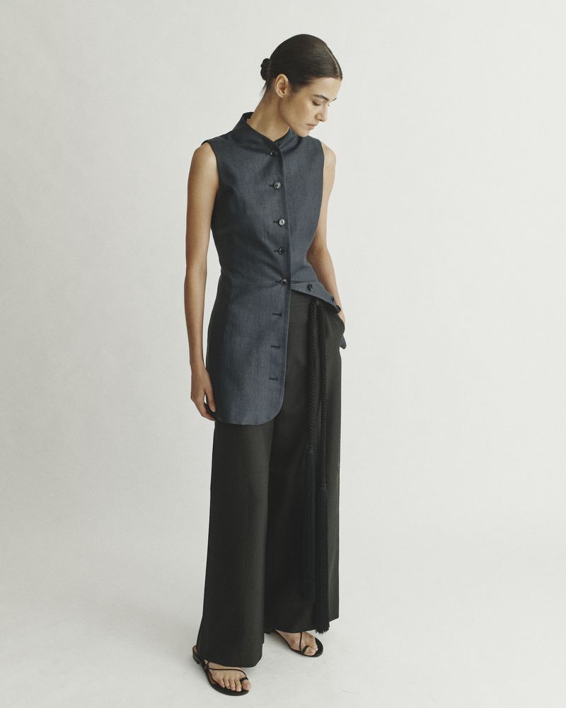 Fashion model poses in a sleeveless navy denim top with an asymmetrical hem and wide-leg black pants in a studio setting.