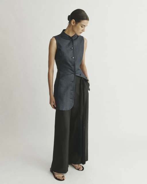 Fashion model poses in a sleeveless navy denim top with an asymmetrical hem and wide-leg black pants in a studio setting.