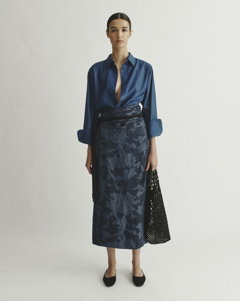 Studio shot of a woman in a blue button-down shirt and a floral pattern skirt, holding a black woven bag against a white backdrop.