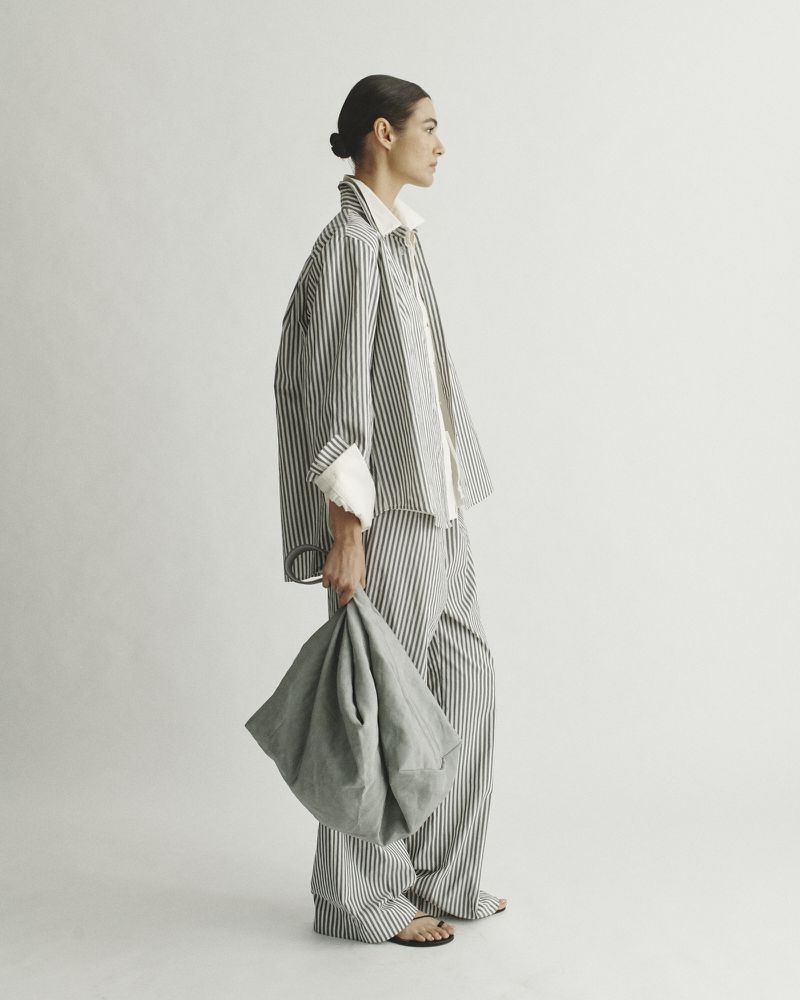 Adult female in striped pajama-style outfit holding a soft bag, side profile in a brightly lit photo studio. Fashion editorial content.