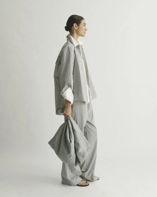 Adult female in striped pajama-style outfit holding a soft bag, side profile in a brightly lit photo studio. Fashion editorial content.